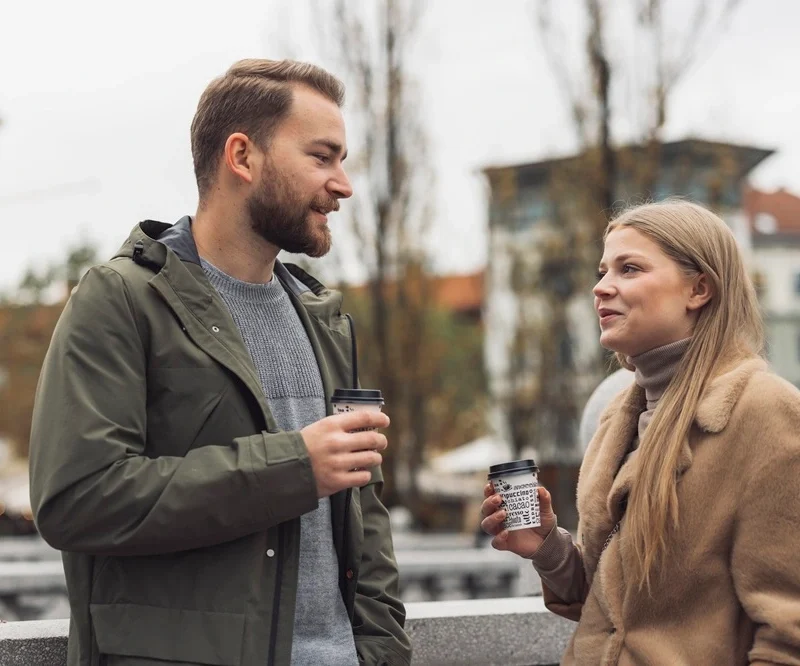 Man and woman chatting outdoors while holding takeaway coffee cups with black lids.